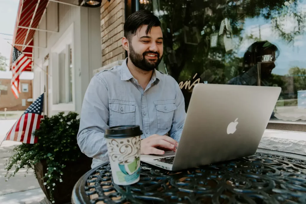 A person is sitting outside at a black metal table with a laptop, smiling. A to-go coffee cup rests on the table. The setting is outdoors, with an American flag and plants visible in the background.