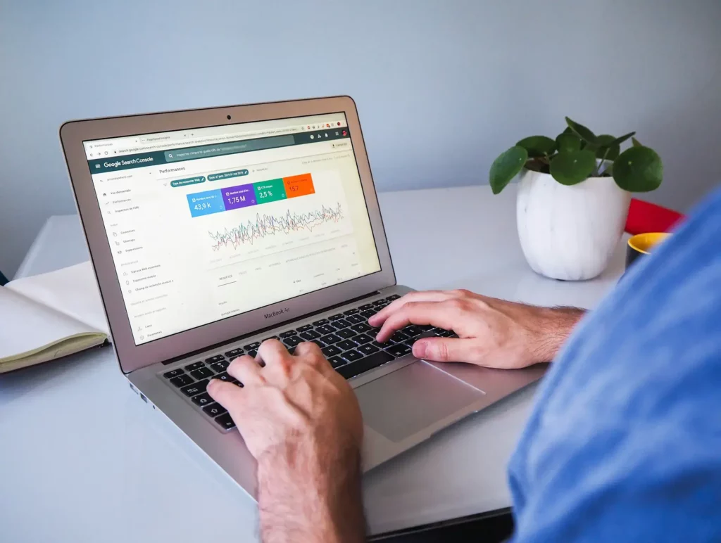 A person types on a laptop displaying a Google Search Console analytics page filled with charts and data. The laptop, nestled next to a small potted plant on the white desk, hints at their unlimited graphic design potential unlocked through a savvy design subscription.