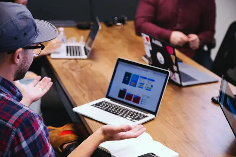 A group of people sit around a wooden table, each with a laptop. The person in the foreground, wearing a cap and glasses, gestures towards their screen, which displays colorful graphs. Casual office setting with focus on teamwork.