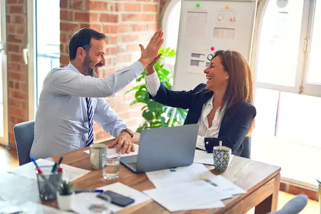 Two colleagues, a man and a woman, are sitting at a wooden desk cluttered with papers. They are smiling and giving each other a high-five. A laptop and a coffee cup are in front of them, with a window and charts in the background.